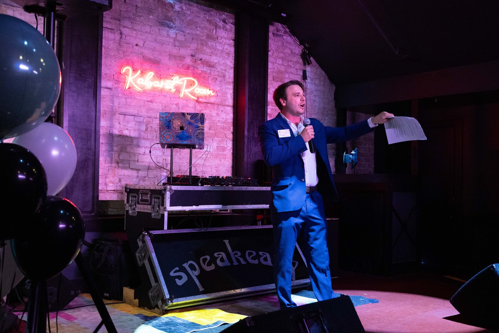 Eric Davis speaking into a microphone on stage under the Speakeasy and Kabaret Room neon signs