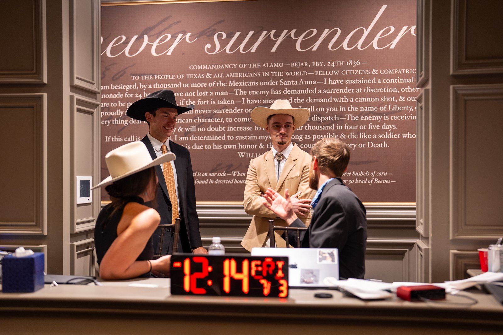 Two hackathon competitors in cowboy hats in front of the Alamo 'never surrender' quote wall