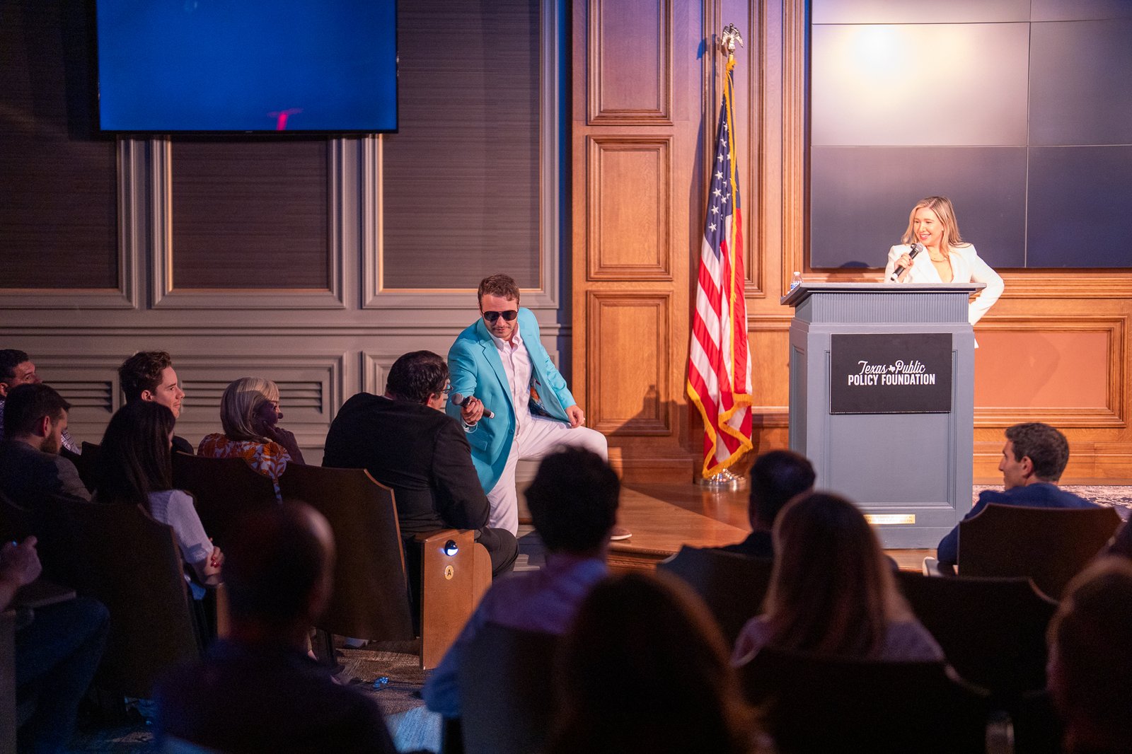 A competitor in sunglasses and a teal blazer striking a pose on stage next to the emcee