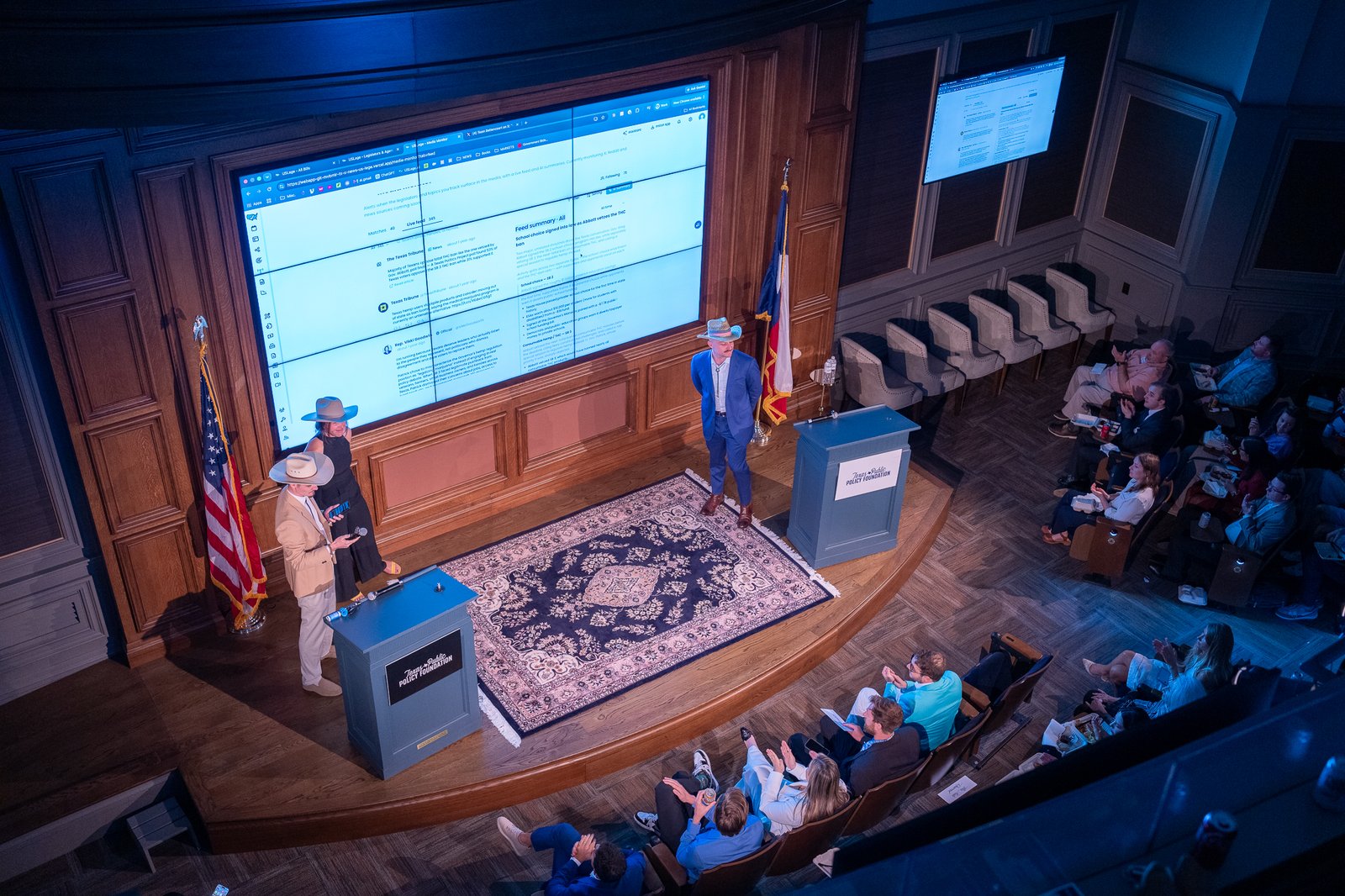 Wide overhead view of the Texas Hackathon audience and stage screens from the balcony